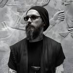 A person with a beard, wearing a beanie, sunglasses, and a dark shirt, standing in front of a carved stone wall.
