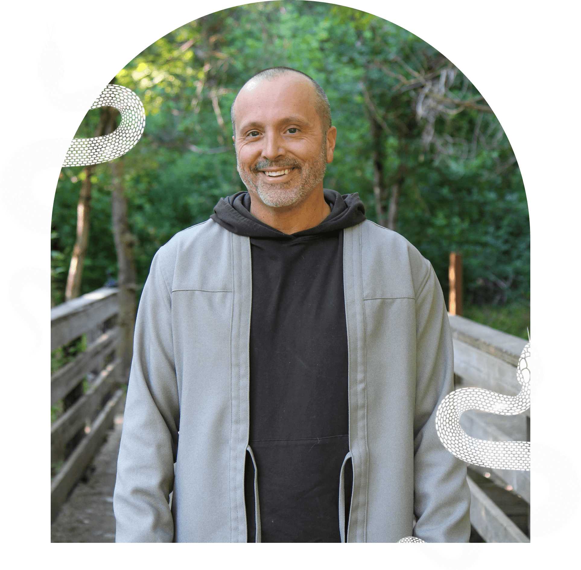 A man with a shaved head, wearing a gray jacket over a black hoodie, stands on a wooden bridge in a forest. Two illustrated snakes are displayed on either side of the image.