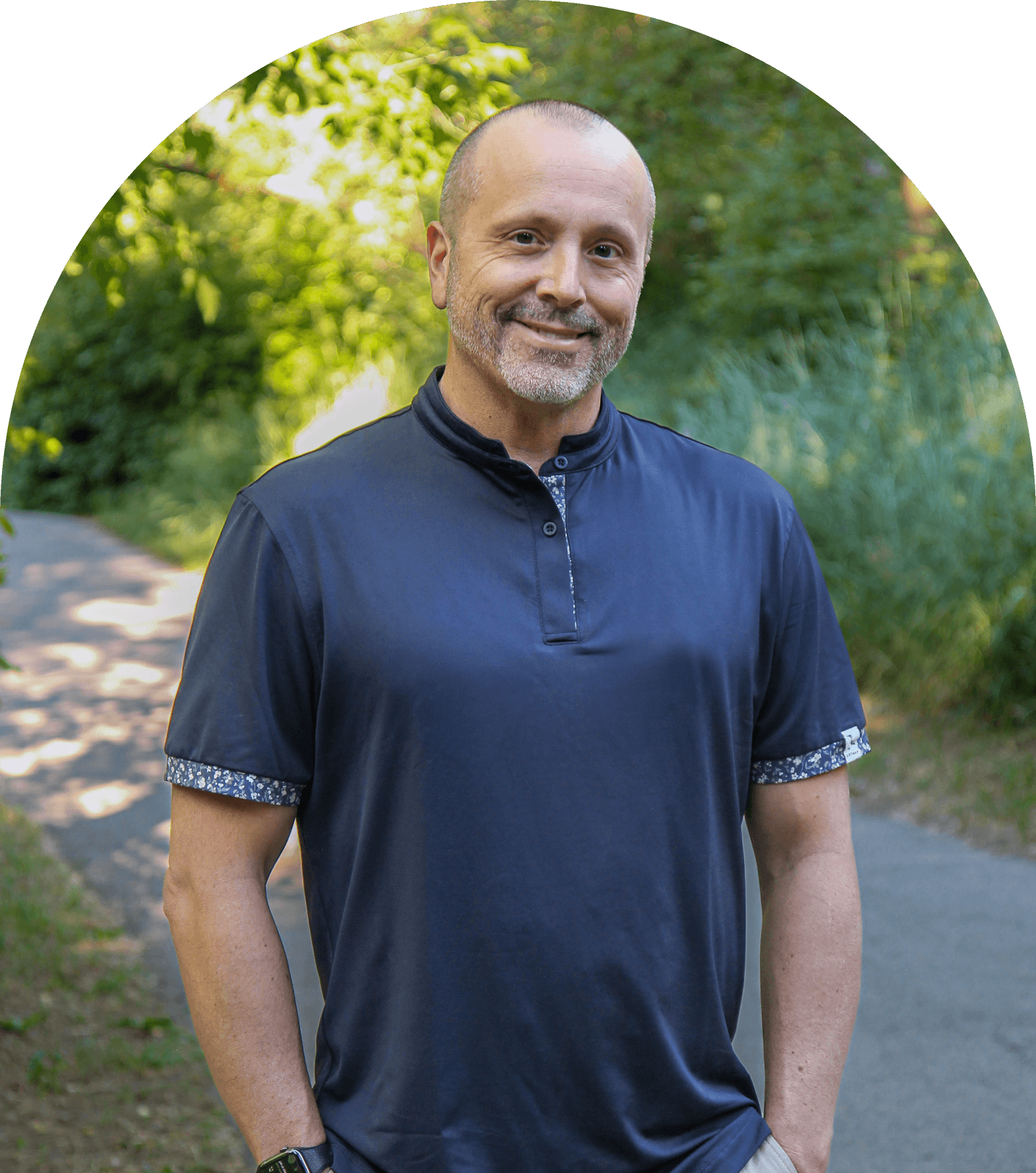 A man with a shaved head and beard smiles while standing on a path surrounded by greenery, wearing a navy blue shirt with patterned cuffs.